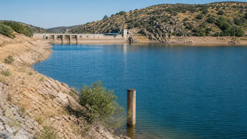 Embalse serrano genérico y vacío de personas con presa, orilla de verano y una regla de nivel sin cifras legibles