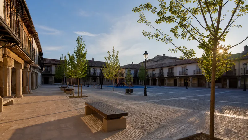 Plaza mayor genérica renovada con soportales, pavimento accesible, bancos, árboles y juegos infantiles