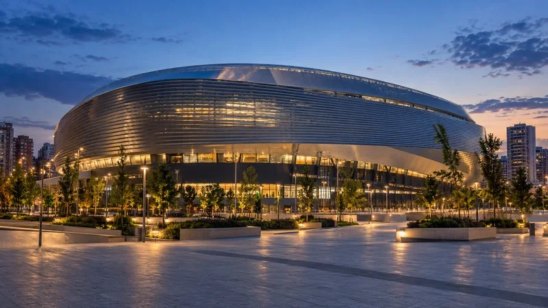 Un estadio de fútbol moderno con fachada metálica iluminada al anochecer y una plaza urbana despejada en primer plano.
